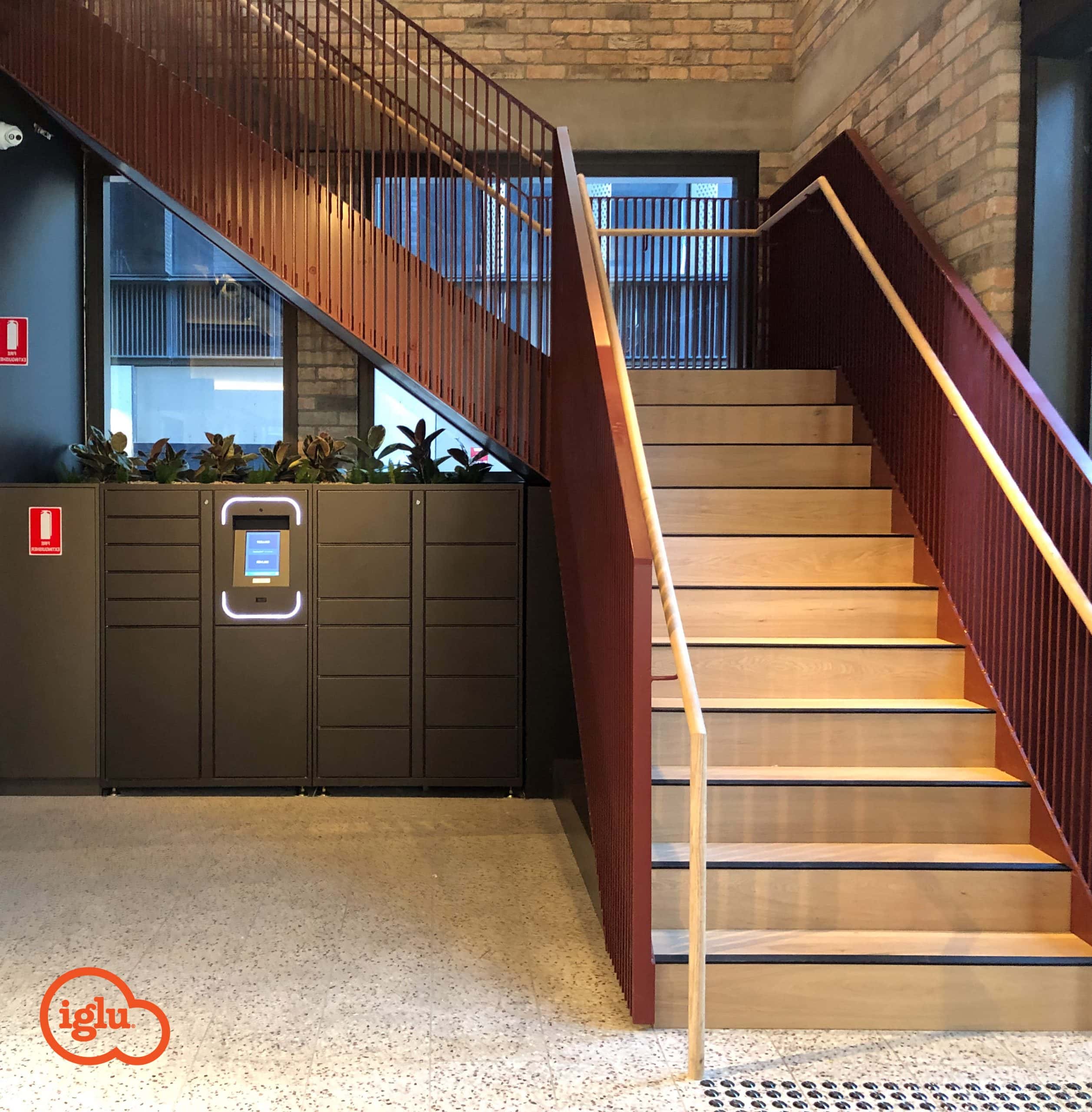 Interior of a modern building featuring a wooden staircase with red railings, parcel management lockers, and a recycling bin under the stairs.