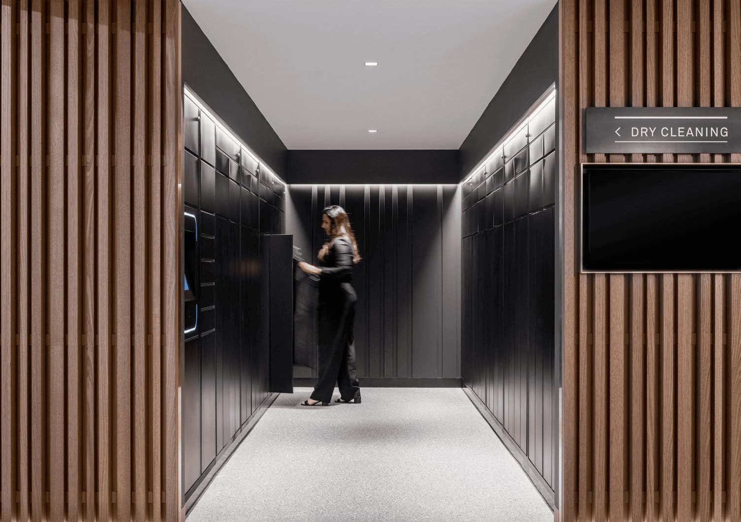 A person in black clothing operates a machine in a sleek, modern hallway with dark walls and wooden paneling. A sign indicates the presence of a dry cleaning service.