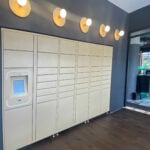 A wall of beige parcel lockers in Perth with a touchscreen panel on the left, illuminated by five round wall lights above, in a modern interior space.