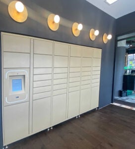 A wall of beige parcel lockers in Perth with a touchscreen panel on the left, illuminated by five round wall lights above, in a modern interior space.